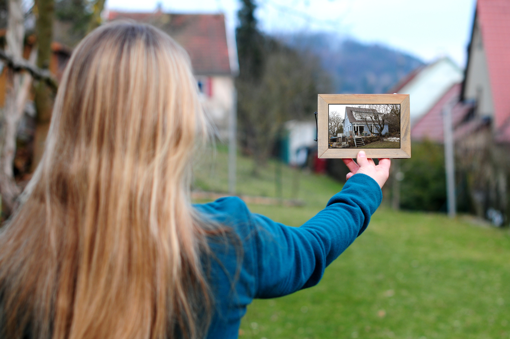 Frau hält einen Bilderrahmen mit einem Foto von einem Haus vor eine Baulücke